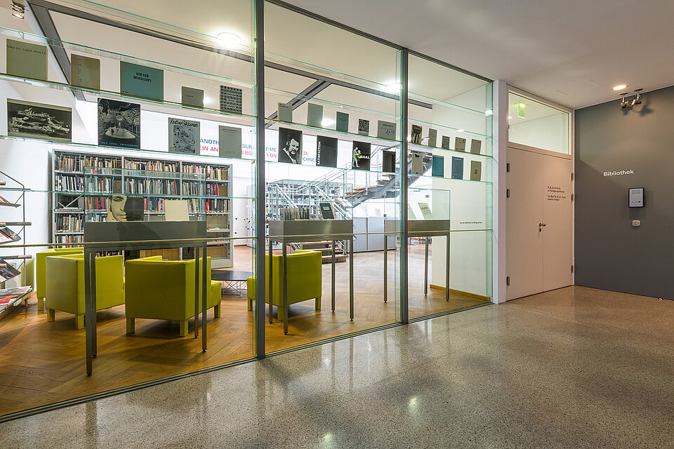 View from the outside into the mumok library, through a glass wall you can see books and a light green seating area