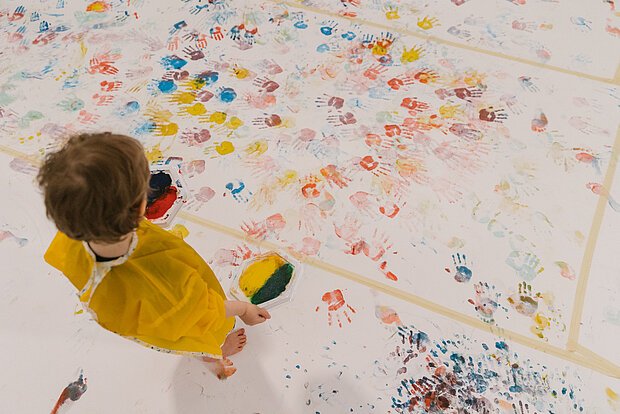
            
                Ein Kind von oben fotografiert, es trägt einen gelben Kittel und steht auf einem weißen Untergrund, der voll mit bunten Farbklecksen ist
            
        