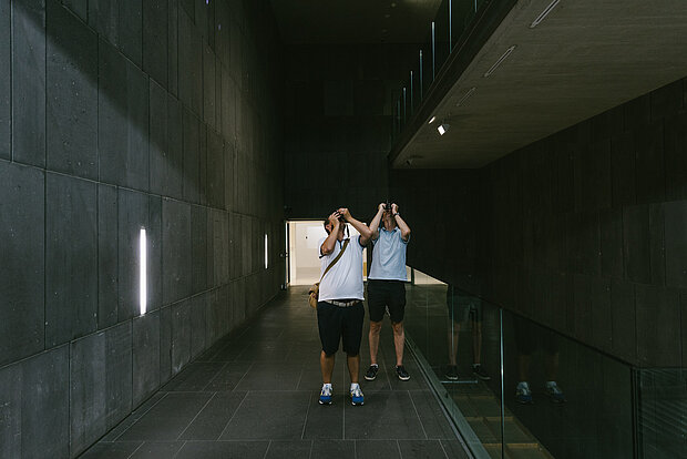 
            
                zwei Personen stehen im mumok Treppenhaus, das mit dunkelgrauen Lavasteinblöcken ausgekleidet ist. Die Personen halten Kameras oder Handys nach oben und scheinen etwas zu fotografieren
            
        