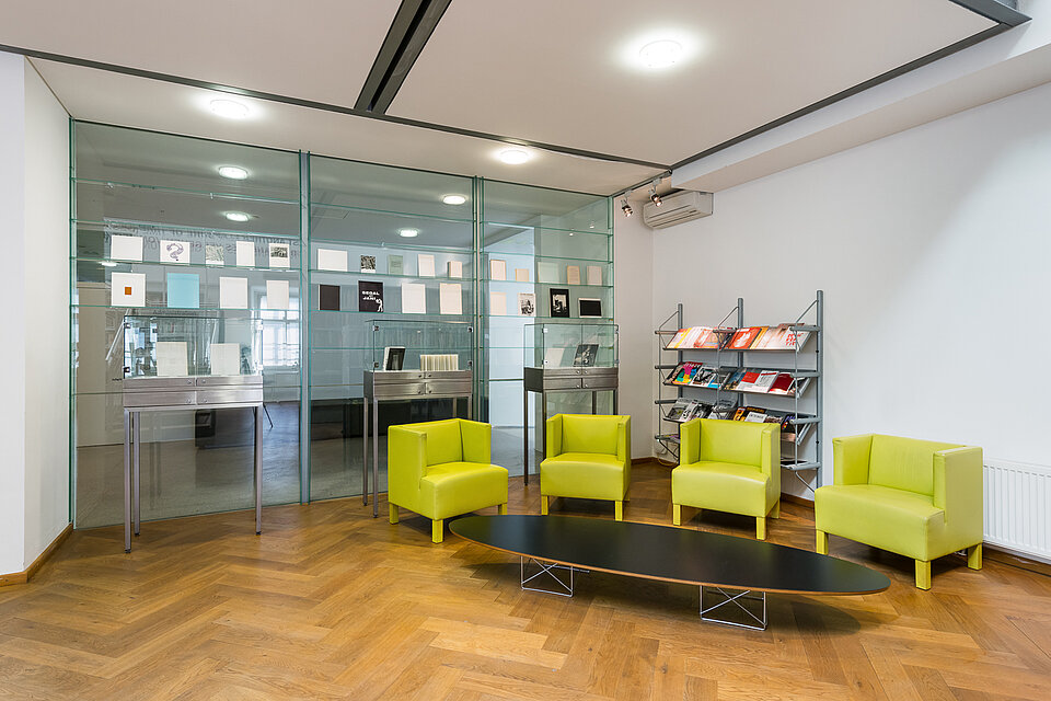 Sitting area in the mumok library, light green armchairs grouped around a low table, a glass wall in the background and book displays in front of it