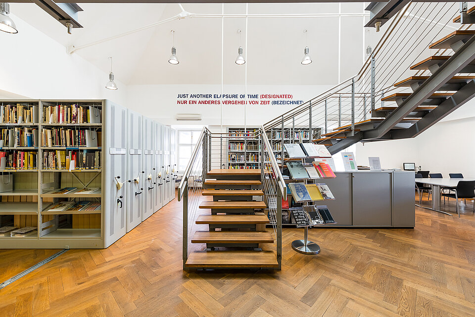 Blick in die mumok Bibliothek, links große Archivschränke mit Büchern, mittig eine Treppe ins Obergeschoss, rechts graue Schränke und Sitzgelegenheiten