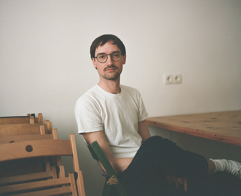 Philipp Fleishcmann sitzend an einem Tisch vor einer weißen Wand, er trägt ein weißes Tshirt und eine Brille