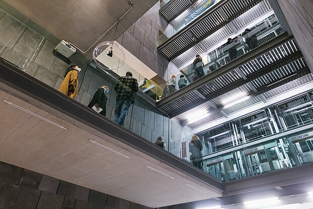 
            
                Blick von unten ins mumok Treppenhaus und auf die Aufzüge, es sind mehrere Ebenen zu sehen, auf denen Menschen stehen
            
        