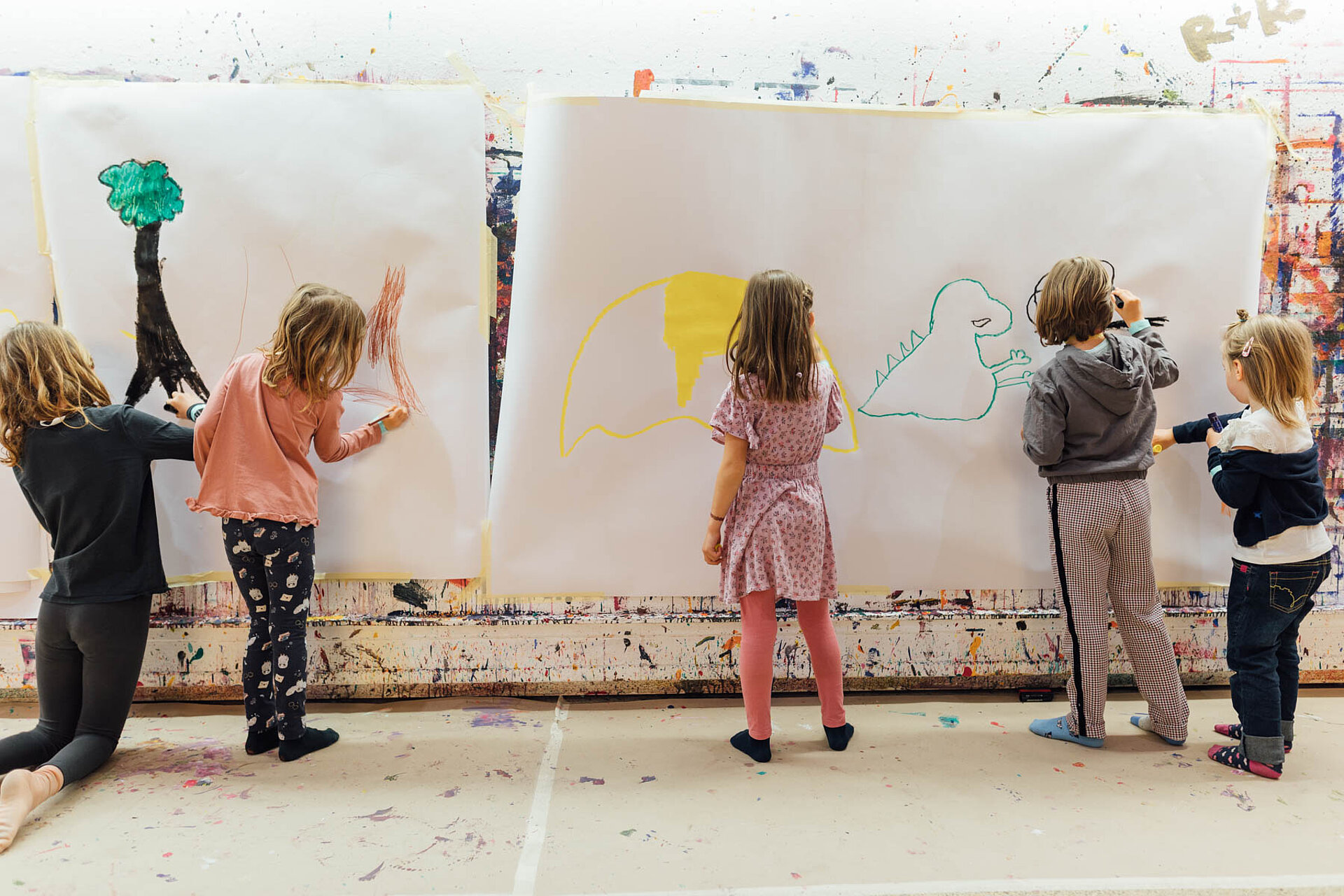 
            
                Kinder malen bunte Motive auf weißes Papier, das an der Wand hängt
            
        