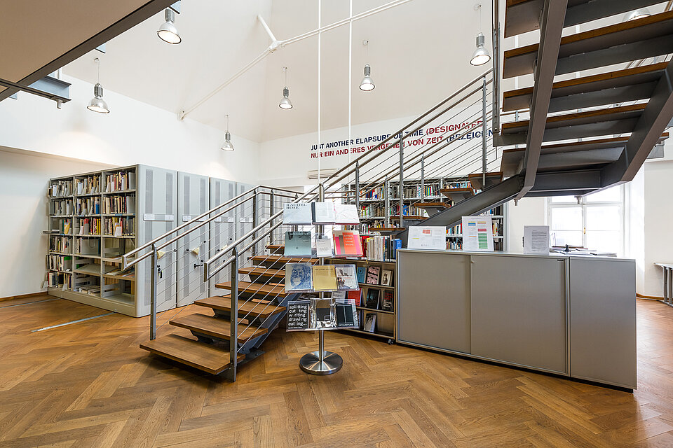 View into the mumok library, on the left are large archive cabinets with books, in the middle a staircase leads up, on the right a gray cabinet