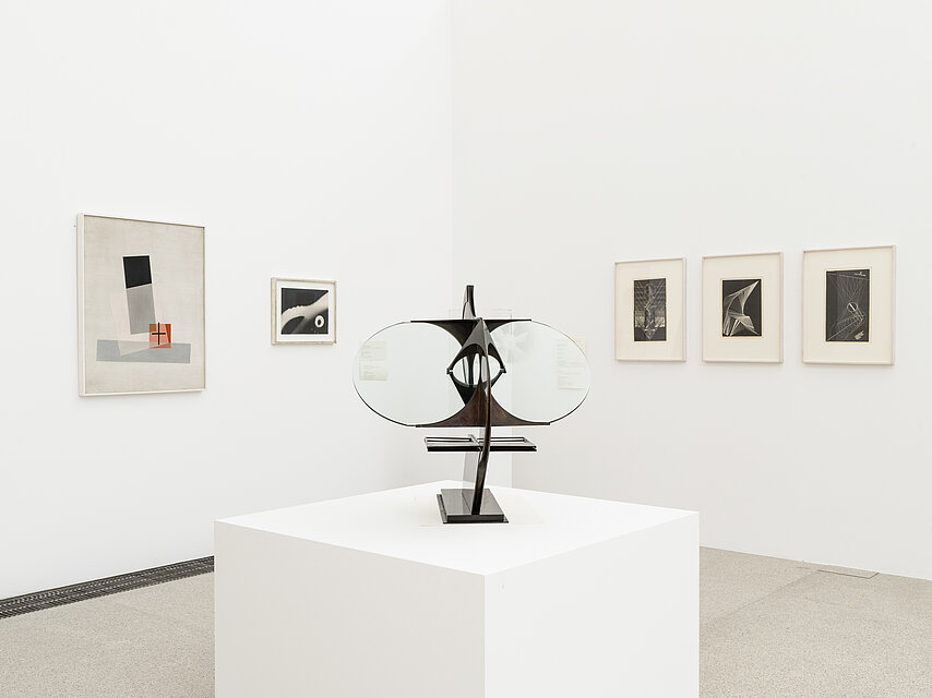 a black, filigree sculpture stands on a white base in the centre of the picture, with pictures of different sizes in dark colours on white walls in the background