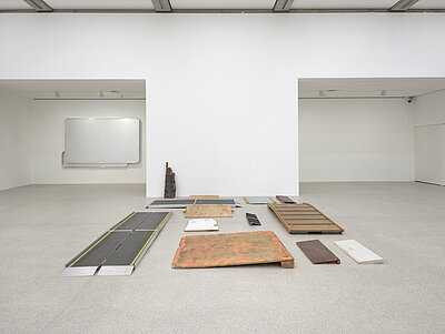 [ID: A collection of wooden and metal ramps arranged on the gallery’s floor in a structured yet fragmented manner. The materials show signs of wear, with some featuring chipped paint and rust. A wall-mounted artwork composed of aluminum panels made from Austrian highway signs, blank yet retaining their original format, is mounted to an alcove wall in the background.]