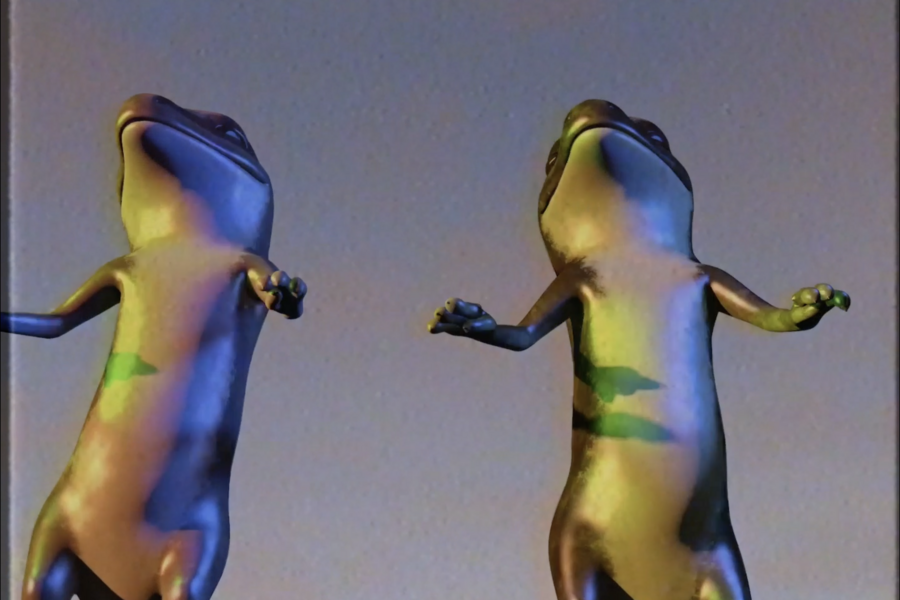 Two upright, stylized frog figures stand side by side, illuminated by colored light. Their smooth bodies display shifting green, blue, and yellow tones against a neutral background.