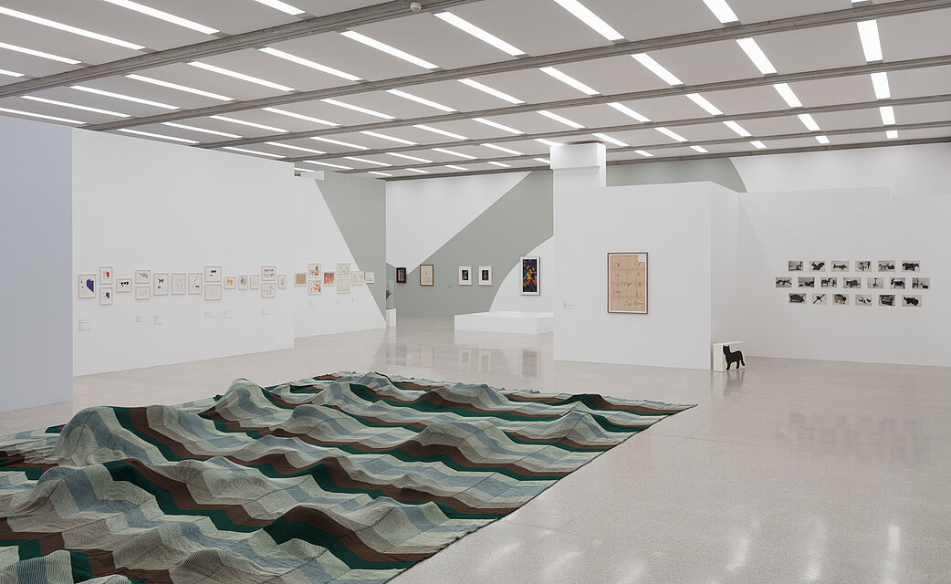  A large-format fabric with a green and blue wave pattern lies on the exhibition floor. Beneath it are invisible stuffed animals that make the fabric appear undulating. On the walls behind it are various pictures in different sizes. 