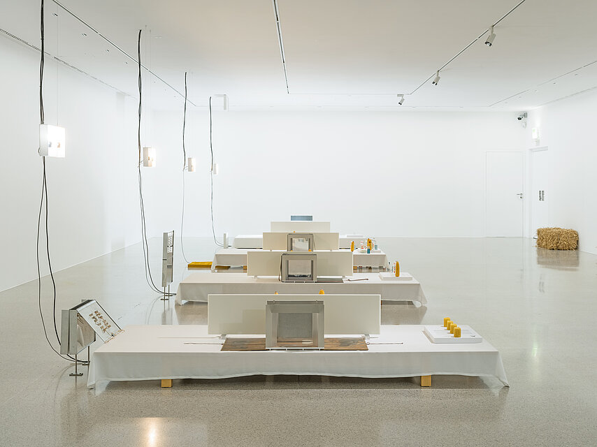 Four white pedestals are lined up one behind the other in the exhibition space. Various sculptures with corncobs are placed on them. A bale of straw stands on the floor in the background.