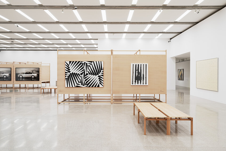 A bright exhibition space with architecture made of light-coloured wood, with abstract, black and white works of art hanging on the wooden displays