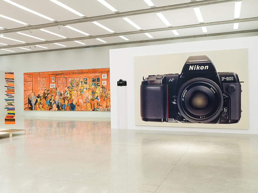Ausstellungsraum mit weißen Wänden und hellem Boden, links im Hintergrund ein orangefarbenes, buntes Gemälde mit vielen Personen, rechts ein großes Bild einer Kamera
