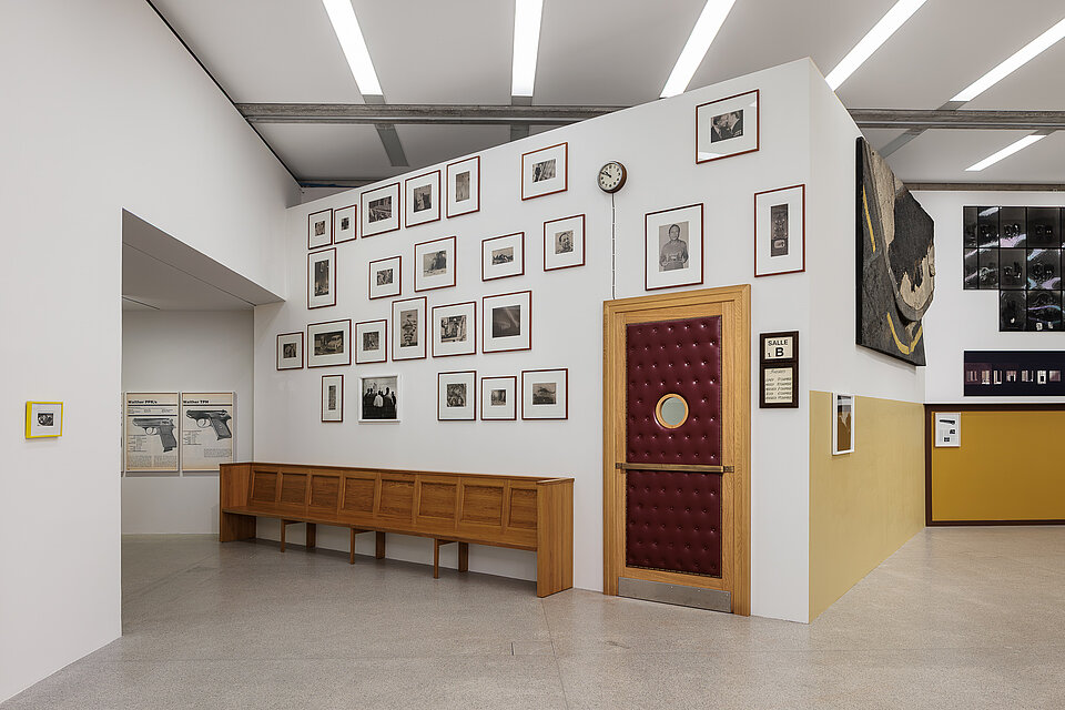 
            
                Ausstellungsansicht mit einer gepolsterten roten Tür, einer langen Holzbank und einer dicht gehängten Gruppe gerahmter Fotografien
            
        