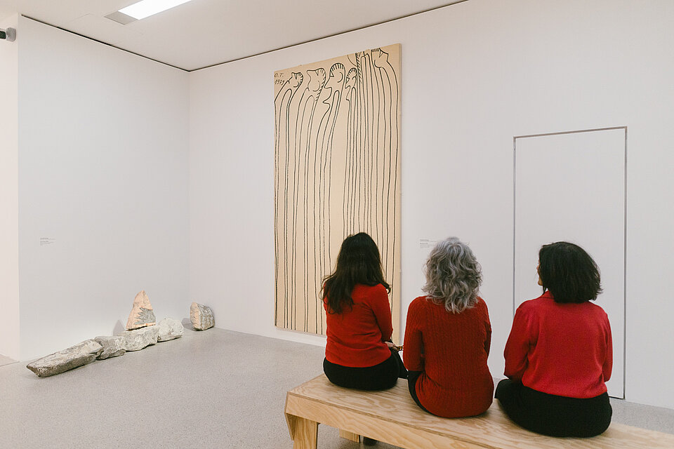 Drei Personen in roten Oberteilen sitzen auf einer Holzbank und betrachten ein hochformatiges Kunstwerk mit vertikalen Linien an einer weißen Museumswand; links liegen mehrere Steinfragmente auf dem Boden. / Three people wearing red tops sit on a wooden bench facing a tall artwork with vertical lines on a white museum wall; several stone fragments are placed on the floor to the left.