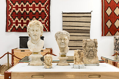 Ein Ausstellungsraum mit weißen Wänden, indem sich zerntral ein große Transportkiste mit darauf platzierten Büsten befindet. Im Hintergrund hängen drei Wandteppiche nebeneinander.