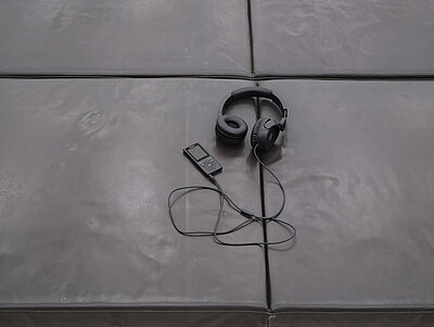 [ID: A pair of headphones connected to a portable audio device lie on a grid of dark leather bench cushions.]