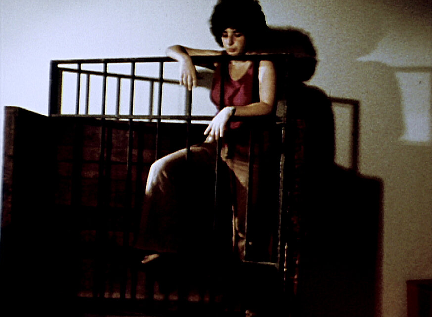 Körniger Videostill einer Person, die auf einer erhöhten Plattform hinter einem schwarzen Metallgeländer sitzt und die Arme auf die obere Stange legt; die Szene ist schwach beleuchtet und wirft deutliche Schatten auf die Wand im Hintergrund. / Grainy video still showing a person seated on a raised platform behind a black metal railing, resting both arms on the top bar; the scene is dimly lit and casts pronounced shadows onto the wall behind.