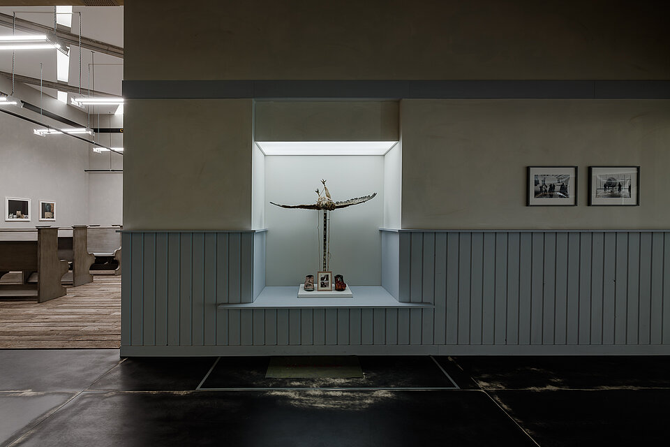 
            
                Wandnische mit beleuchteter Vitrine, die eine skulpturale vogelähnliche Form mit ausgebreiteten Flügeln und kleinere Objekte auf einem Sockel zeigt; rechts daneben hängen zwei gerahmte Fotografien.
            
        