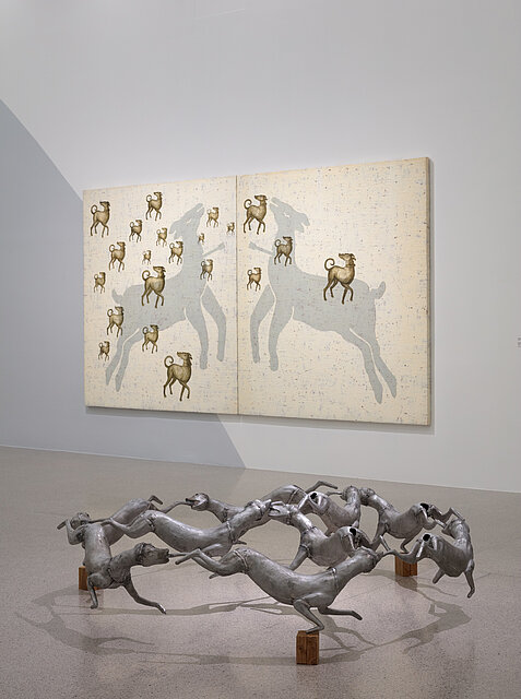Two beige pictures of dogs hang in the background. In the foreground an abstract sculpture of metal dogs.