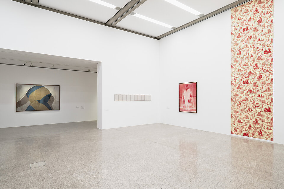 ein heller Ausstellungsraum, links gibt es eine Art Nische, in der ein abstraktes Bild hängt. Rechts hängt von der Decke bis zum Boden eine große textile Arbeit in Rot und Gelb und Weiß, links daneben ein rotes Gemälde