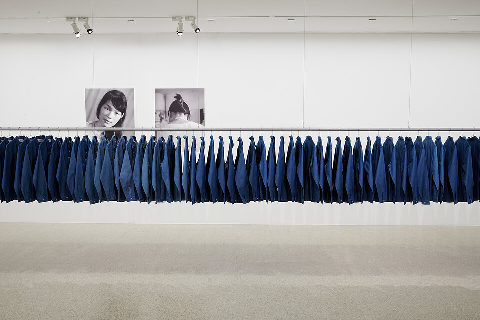 lange Kleiderstange mit blauen Hemden, im Hintergrund schwarz weiße Portraits, heller Ausstellungsraum