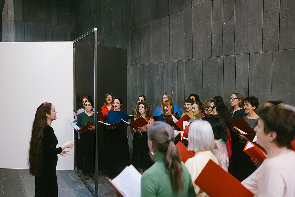 Gruppe von Personen mit geöffneten roten Chormappen steht in einem Ausstellungsraum mit dunklen Betonwänden; im Vordergrund dirigiert eine Person in dunkler Kleidung vor einem mobilen Trennelement. / Group of people holding open red choir folders stands in an exhibition space with dark concrete walls; in the foreground, a person in dark clothing conducts in front of a movable partition.
