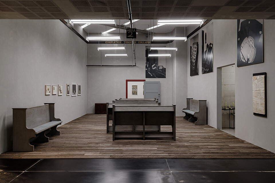 
            
                Ausstellungsraum mit holzverkleidetem Boden und mehreren bankähnlichen Sitzreihen, an den Wänden hängen Fotografien und großformatige schwarz-weiße Bildarbeiten.
            
        