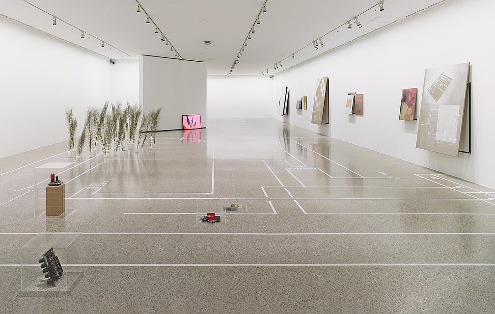 Dried grasses stand in the exhibition room, a screen with a red image can be seen in the background 