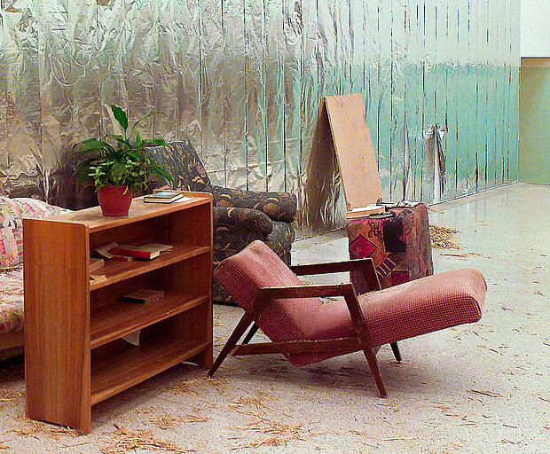 
            
                Installation featuring living room furniture in an exhibition space: a low wooden bookshelf with a potted plant and books, an upholstered lounge chair, a sofa, and a suitcase are arranged in front of a large reflective wall. Wood shavings and scattered debris on the floor contribute to the appearance of a temporary or artistic setting.
            
        