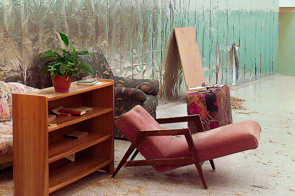 Installation mit Wohnzimmermöbeln in einem Ausstellungsraum: Ein niedriges Holzregal mit Topfpflanze und Büchern, ein gepolsterter Sessel, ein Sofa und ein Koffer stehen vor einer großflächigen Wand aus reflektierendem Material. Auf dem Boden liegen Holzspäne und weitere Spuren eines provisorischen oder künstlerischen Arrangements.