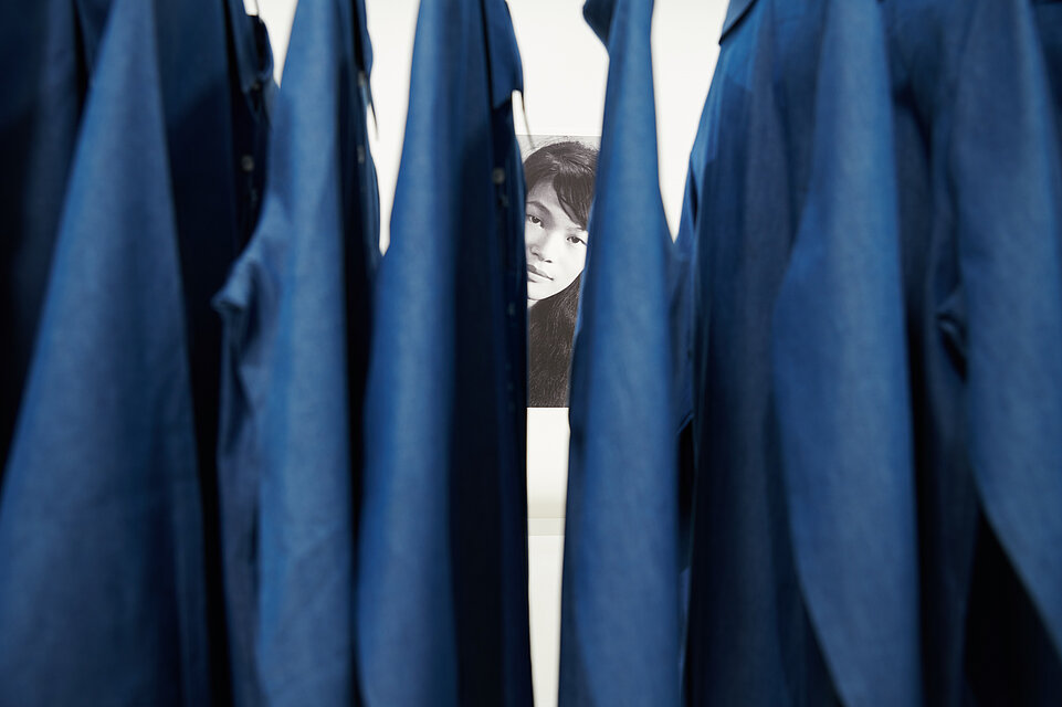 Blue items of clothing hang on a pole, a black and white portrait photo of a woman flashes out through a gap