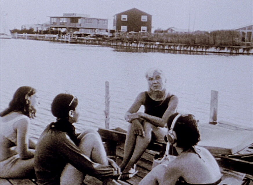 Schwarz-weißes Filmbild einer Gruppe von vier Personen auf einem Steg oder einer Plattform am Wasser; im Hintergrund sind Uferbebauung, Schilf und mehrere Gebäude zu sehen. / Black-and-white film still showing a group of four people seated on a dock or platform by the water; buildings, reeds, and shoreline structures are visible in the background.