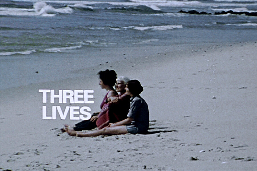 Videostill mit drei Personen, die nebeneinander am Sandstrand sitzen und auf das Meer blicken; links daneben steht der weiße Schriftzug „THREE LIVES“. / Film still showing three people seated side by side on a sandy beach facing the ocean, with the white text “THREE LIVES” displayed beside them.
