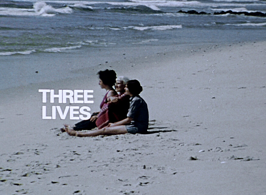 Videostill mit drei Personen, die nebeneinander am Sandstrand sitzen und auf das Meer blicken; links daneben steht der weiße Schriftzug „THREE LIVES“. / Film still showing three people seated side by side on a sandy beach facing the ocean, with the white text “THREE LIVES” displayed beside them.