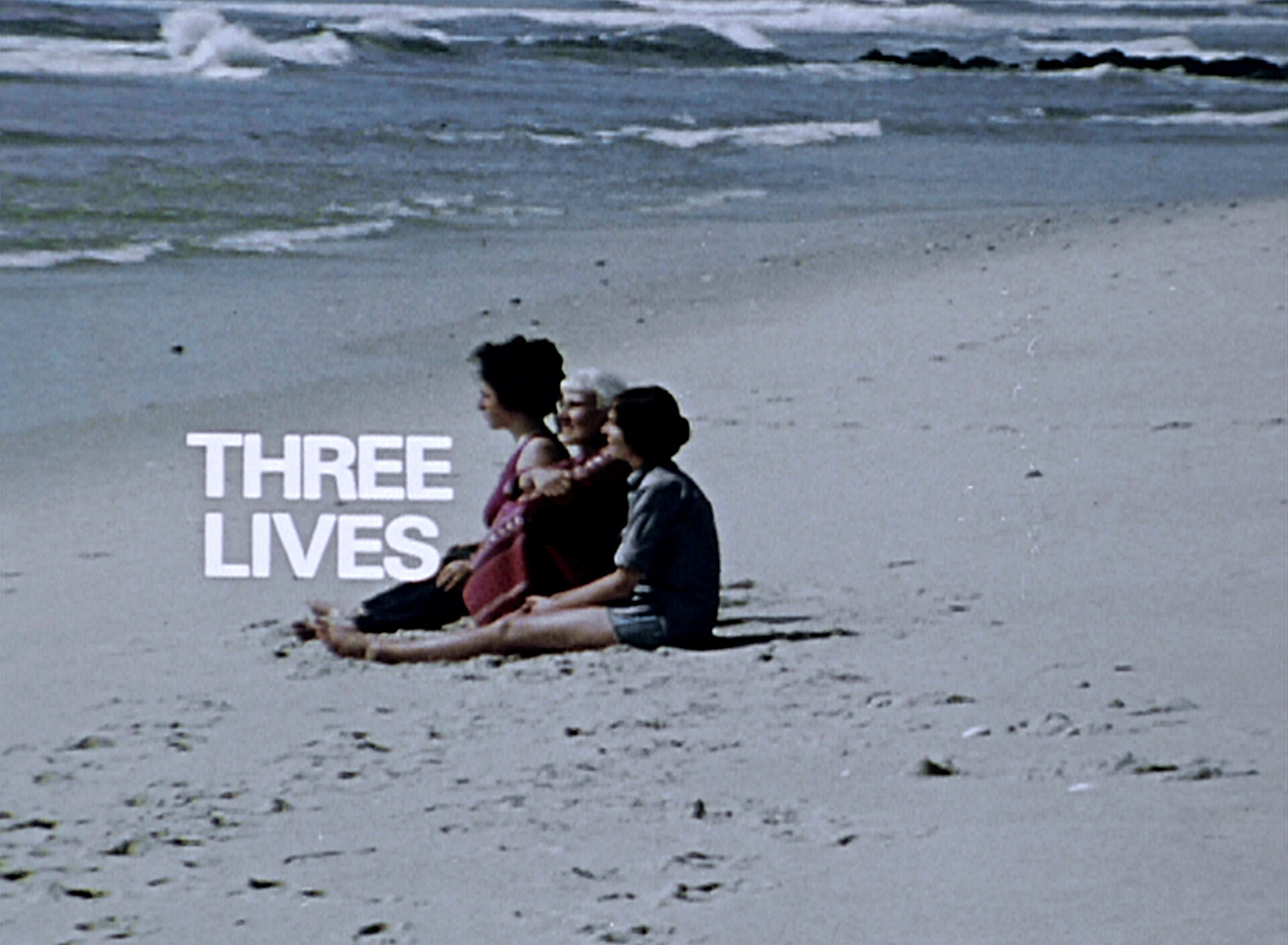 Videostill mit drei Personen, die nebeneinander am Sandstrand sitzen und auf das Meer blicken; links daneben steht der weiße Schriftzug „THREE LIVES“. / Film still showing three people seated side by side on a sandy beach facing the ocean, with the white text “THREE LIVES” displayed beside them.