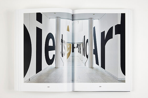 
            
                Aufgeschlagenes Buch vor weißem Hintergrund mit einer doppelseitigen Fotografie eines hellen Innenraums, dessen Wände von übergroßen schwarzen Buchstaben und typografischen Elementen bedeckt sind. / Open book against a white background showing a double-page photograph of a bright interior space whose walls are covered with oversized black letters and typographic elements.
            
        