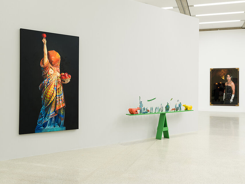 Kunstwerke in einem hellen Ausstellungsraum, links eine Frau, die in bunte Tücher gehüllt wie die Freiheitsstatue posiert, gemalt auf schwarzem Grund