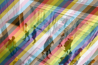 Blick ins Treppenhaus des mumok mit verglasten Fahrstühlen, es gehen mehrere Menschen auf unterschiedlichen Ebenen herum. Das Bild ist mit einem transparenten Regenbogen versehen.