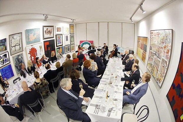 
            
                Foto einer größeren Gruppe an Menschen, die an zwei langen Tafeln angeordnet sitzen. An den Wänden hängen Kunstwerke.
            
        