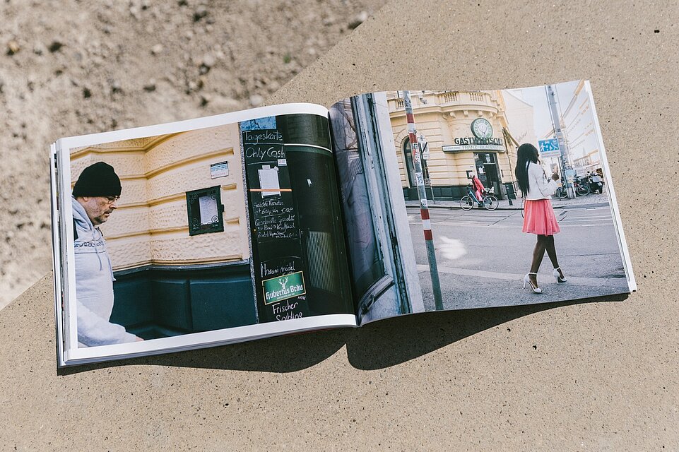 Aufgeschlagenes Fotobuch auf einer Betonfläche mit einer Doppelseite urbaner Straßenszenen, links eine Person neben einer Gebäudefassade und Hinweistafel, rechts eine Person an einer Straßenkreuzung mit historischen Gebäuden im Hintergrund. / An open photo book placed on a concrete surface showing a double-page spread of urban street scenes, with a person next to a building facade and notice board on the left and another person at a street intersection with historic buildings in the background on the right.