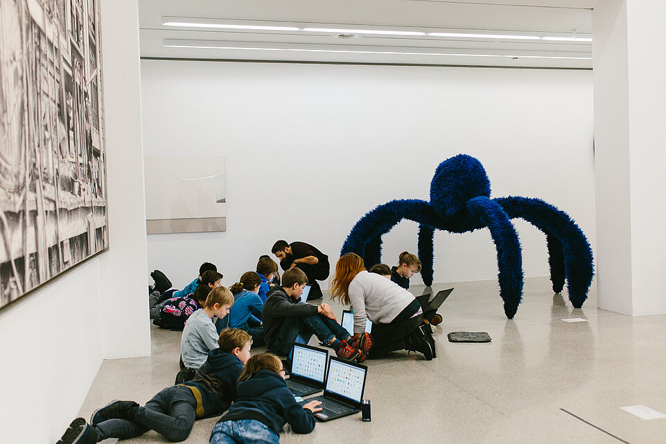 Children sit on the floor in the museum and work on laptops, in the background a large blue sculpture of a plush spider