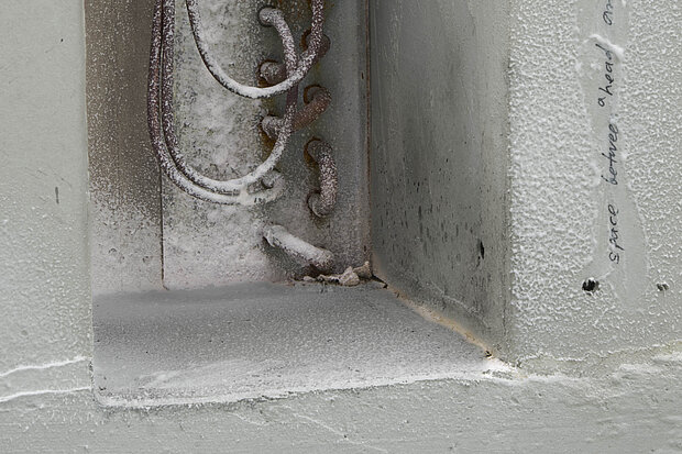 
            
                Detailansicht einer wandartigen Nische mit rauer, weißer Oberfläche, sichtbar eingegossenen Metallbügeln und Spuren von Staub, Abrieb und handschriftlichen Markierungen. / Close-up view of a wall-like recess with a rough white surface, embedded metal loops, and visible traces of dust, abrasion, and handwritten markings.
            
        