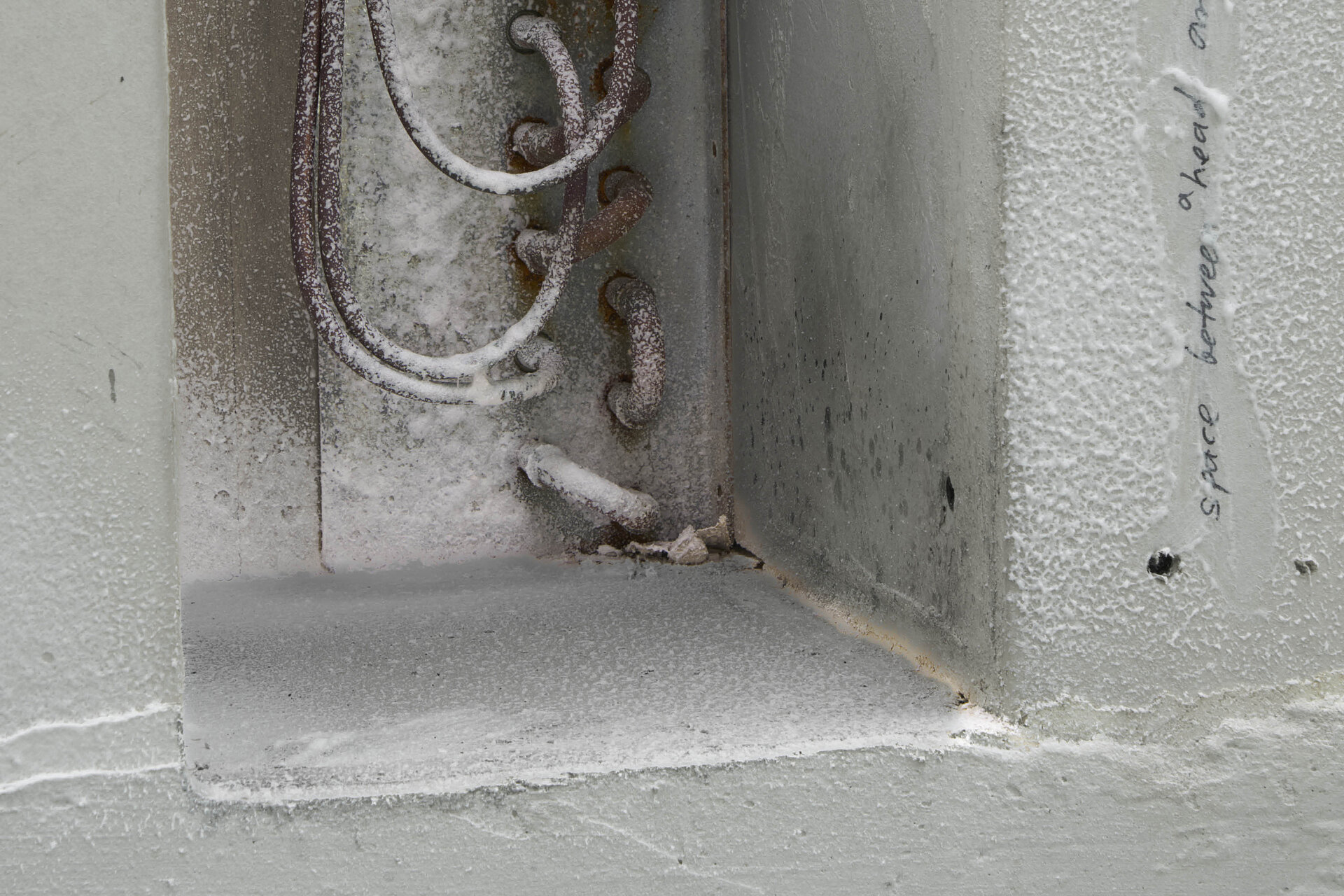 Detailansicht einer wandartigen Nische mit rauer, weißer Oberfläche, sichtbar eingegossenen Metallbügeln und Spuren von Staub, Abrieb und handschriftlichen Markierungen. / Close-up view of a wall-like recess with a rough white surface, embedded metal loops, and visible traces of dust, abrasion, and handwritten markings.
