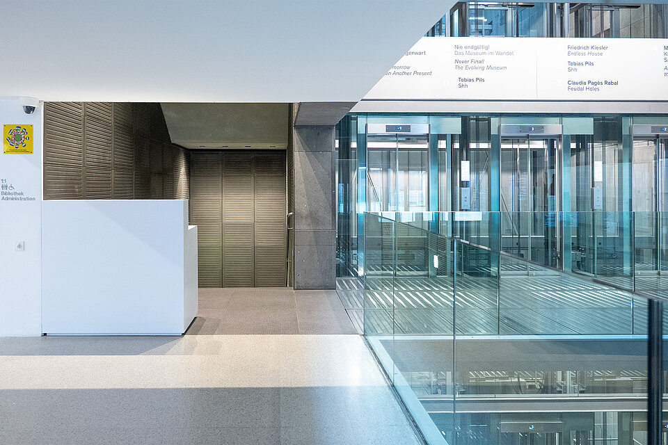 Moderner Innenbereich mit Glasgeländer und Blick auf mehrere Aufzüge, links eine weiße Empfangstheke vor einer strukturierten Wand und Beschilderung im oberen Bereich. / Modern interior space with a glass railing overlooking multiple elevators, with a white reception desk on the left in front of a textured wall and signage above.