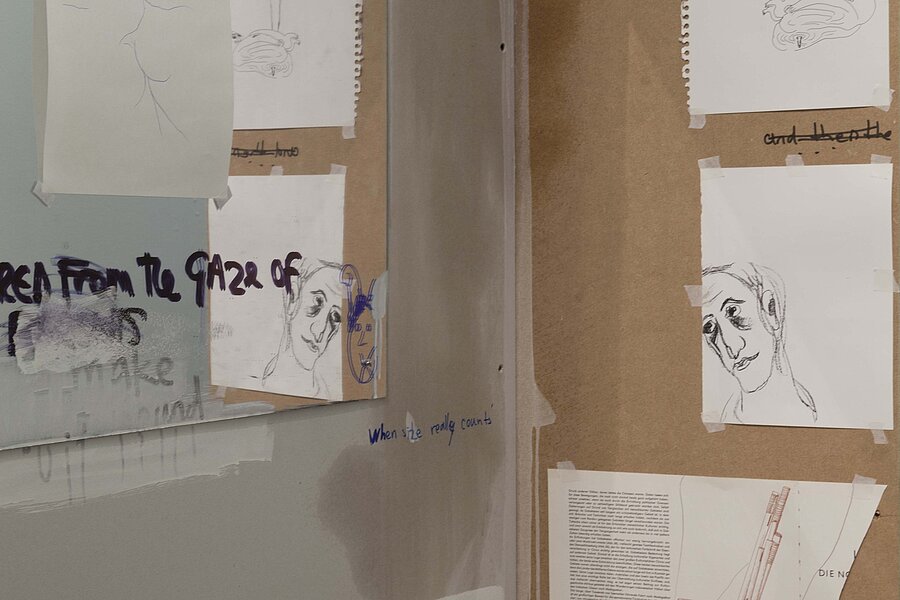 Atelier- oder Ausstellungswand mit mehreren aufgeklebten Papierarbeiten, darunter skizzenhafte Porträtzeichnungen und handschriftliche Notizen auf Karton und Papier. / Studio or exhibition wall with several taped paper works, including sketch-like portrait drawings and handwritten notes on cardboard and paper.