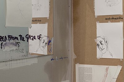 Atelier- oder Ausstellungswand mit mehreren aufgeklebten Papierarbeiten, darunter skizzenhafte Porträtzeichnungen und handschriftliche Notizen auf Karton und Papier. / Studio or exhibition wall with several taped paper works, including sketch-like portrait drawings and handwritten notes on cardboard and paper.