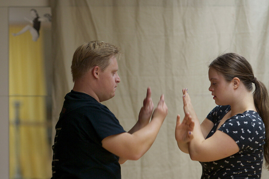 Two people stand facing each other and touch palms with raised hands as part of an interaction or exercise. A beige curtain hangs in the background, and a mirror with a small figure on it is visible on the left.