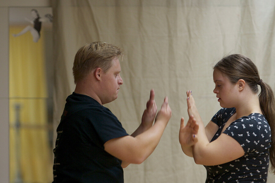 Zwei Personen stehen sich gegenüber und berühren mit erhobenen Händen die Handflächen, als Teil einer Interaktion oder Übung. Im Hintergrund hängt ein beiger Vorhang, links ist ein Spiegel mit einer kleinen Figur darauf sichtbar.
