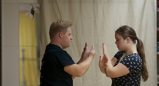 
            
                Zwei Personen stehen sich gegenüber und berühren mit erhobenen Händen die Handflächen, als Teil einer Interaktion oder Übung. Im Hintergrund hängt ein beiger Vorhang, links ist ein Spiegel mit einer kleinen Figur darauf sichtbar.
            
        