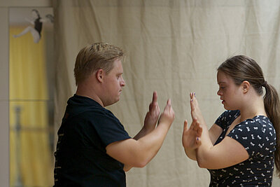 Zwei Personen stehen sich gegenüber und berühren mit erhobenen Händen die Handflächen, als Teil einer Interaktion oder Übung. Im Hintergrund hängt ein beiger Vorhang, links ist ein Spiegel mit einer kleinen Figur darauf sichtbar.
